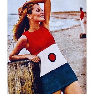 May include: A woman wearing a red, white, and blue mini dress with a large red circle on the white section. She is standing on a wooden pier with the ocean in the background.