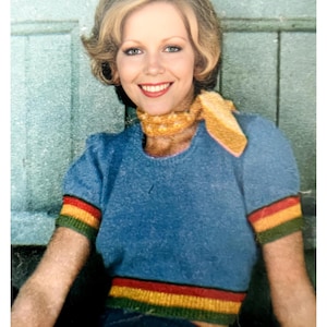 Puede incluir: Un suéter azul de punto con rayas de colores en las mangas y el dobladillo inferior. El suéter tiene un cuello redondo y lo lleva una mujer con el pelo corto y rubio.