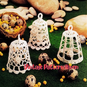 May include: Three white crochet bell ornaments with intricate openwork designs. The bells are displayed with speckled eggs, a woven nest, and small yellow beads on a green surface. The text "Vintage Pattern Haven" is visible.