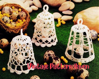 May include: Three white crochet bell ornaments with intricate openwork designs. The bells are displayed with speckled eggs, a woven nest, and small yellow beads on a green surface. The text "Vintage Pattern Haven" is visible.