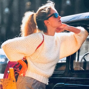 Könnte beinhalten: Cremefarbener Strickpullover mit Rundhalsausschnitt und langen, voluminösen Ärmeln. Der Pullover hat ein strukturiertes Strickmuster und ein roter Riemen ist sichtbar. Die Person trägt eine Sonnenbrille und hat ihr Haar zu einem Pferdeschwanz gebunden.