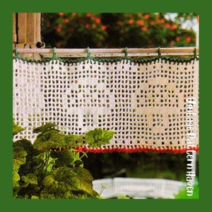 May include: A white crocheted curtain with a square pattern hangs in front of a window. The top edge is trimmed with green, and the bottom edge has a red border. Green foliage is visible in the foreground, with a blurred outdoor background.