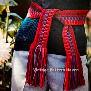 Könnte beinhalten: Ein gewebter Gürtel mit roten und violetten Streifen und Quasten. Der Gürtel ist um die Taille einer Person gebunden.