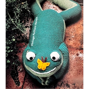 May include: A green knitted frog toy with a yellow butterfly on its nose. The frog is lying on its back with its legs outstretched. The toy is made with a textured knit fabric and has large, white eyes.