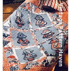 May include: A quilt with a patchwork design featuring appliqued figures of girls wearing bonnets and dresses. The quilt is made with a variety of fabrics in shades of pink, red, blue, and white. The quilt is bordered with a floral print fabric. The text "Vintage Pattern Haven" is printed on the quilt.