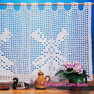 Può includere: Una tenda all'uncinetto bianca con motivo a griglia appesa a un bastone di legno. La tenda presenta un sottile disegno floreale nella trama. Una piccola brocca in ceramica e una pianta in vaso con fiori viola si trovano davanti.