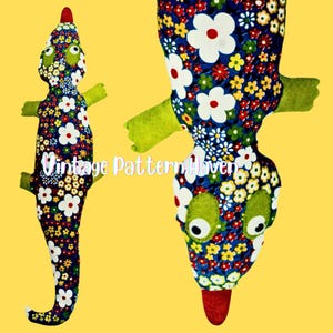 May include: A stuffed lizard toy with a floral pattern in blue, white, red, and yellow. The lizard has green arms and eyes, and a red tip on its nose. The text "Vintage Pattern Haven" is visible on the toy.
