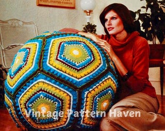 May include: A large, colorful, crocheted, hexagonal-shaped beanbag with a blue, yellow, green, and brown color scheme. The beanbag is sitting on a floor and a woman is sitting next to it.