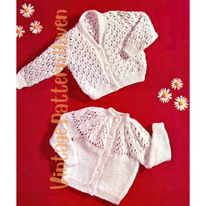 May include: Two white knitted baby cardigans with button closures. The cardigans have a lacy pattern and are laid out on a red background.