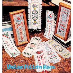 May include: A collection of handmade cross-stitch bookmarks and framed pieces. Designs include houses, pineapples, strawberries, and floral patterns. Text includes "Home", "Cookin'", "Bedtime Stories", and an alphabet. The bookmarks have lace edges.