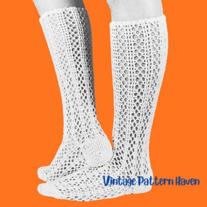Puede incluir: Un par de calcetines blancos hasta la rodilla con un intrincado patrón de ganchillo calado y un diseño de malla hexagonal. Los calcetines se extienden desde el pie hasta la pantorrilla, mostrando una textura de encaje uniforme en toda la prenda.