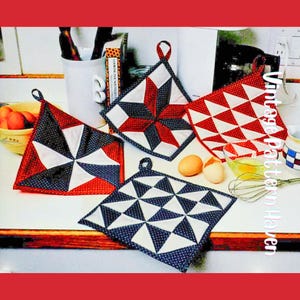 以下が含まれることがあります： 赤、白、ネイビーブルーのキルト鍋つかみのコレクション。鍋つかみは幾何学的な星と三角形のパターンが特徴です。それぞれに吊り下げ用のループが付いています。卵と泡立て器も表示されています。