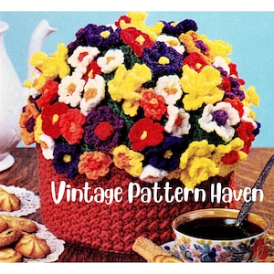 Puede incluir: Calienta-tetera de ganchillo en forma de maceta con una variedad de flores de colores. El calienta-tetera es rojo y las flores son amarillas, naranjas, rojas, moradas y blancas. El texto "Vintage Pattern Haven" está impreso en el calienta-tetera.