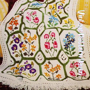 May include: A white crocheted blanket with a floral pattern. The blanket features various colorful flowers, including pink, orange, purple, and blue, set within green hexagonal frames. The border is white with a fringe.