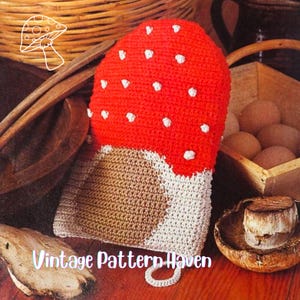 Peut inclure: Un gant de cuisine en forme de champignon crocheté, avec un chapeau rouge et des pois blancs. Le gant a un pied beige et un bord blanc. Le texte "Vintage Pattern Haven" est visible.