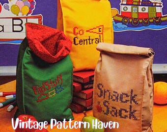 May include: Three fabric lunch bags in yellow, green, and tan, each embroidered with text. The yellow bag says "Co-Central", the green bag says "KidStuff", and the tan bag says "Snack Sack". Apples, pears, and a small snack are in the foreground.