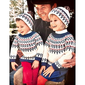 May include: A father and two children wearing matching white, blue, and red patterned sweaters with pom-pom hats. The sweaters have a fair isle design.