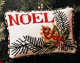 Könnte beinhalten: Ein weißes Kissen mit einem roten gestickten "NOEL" und einem grünen und braunen Kiefernzweig-Design. Das Kissen hat einen schwarz-weißen Muster-Rand.