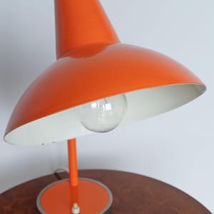 Puede incluir: Una lámpara de escritorio de color naranja brillante con una pantalla interior blanca. La lámpara tiene una base de metal y un diseño sencillo.