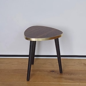 Puede incluir: Una pequeña mesa auxiliar de tres patas con una tapa triangular. La tapa es de color marrón oscuro con un borde dorado. Las patas negras y anguladas crean una estética moderna de mediados de siglo. La mesa está sobre un suelo de madera.