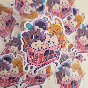 May include: A die-cut sticker featuring a pink shopping cart filled with four cartoon characters: a brown cat, a white fluffy creature, a yellow fox-like figure in a hood, and a purple hooded character with a blue halo, all in a colorful style.