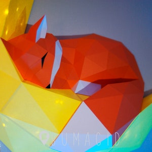 Pu&ograve; includere: Una scultura di volpe in origami rosso con un viso bianco, che dorme su uno sfondo di origami giallo e verde. Lo sfondo ha la scritta "BUMAGID" stampata.