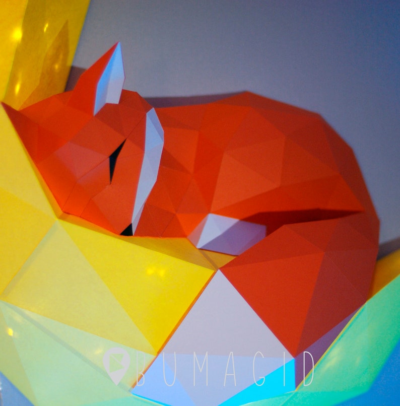 Pu&ograve; includere: Una scultura di volpe in origami rosso con un viso bianco, che dorme su uno sfondo di origami giallo e verde. Lo sfondo ha la scritta "BUMAGID" stampata.