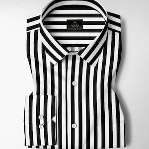 Puede incluir: Camisa a rayas blancas y negras doblada con cuello clásico y puños abotonados. La camisa presenta rayas verticales y una pequeña etiqueta que dice "KOLLER CUT". La camisa está hecha de una tela crujiente, probablemente de algodón.