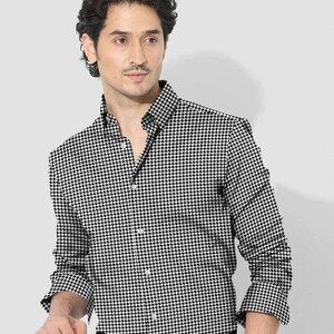 Pode incluir: Uma camisa abotoada com padr&atilde;o pied-de-poule em preto e branco. A camisa de manga comprida tem uma gola cl&aacute;ssica e &eacute; abotoada na frente. A camisa &eacute; combinada com cal&ccedil;as pretas.