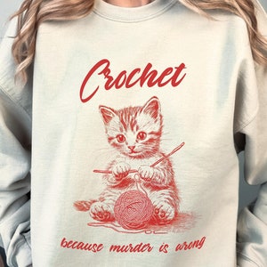 May include: A light beige sweatshirt with the text "Crochet because murder is wrong" in red. A cartoon image of a cat crocheting with yarn is above the text.