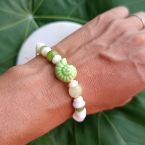 May include: A bracelet with a light green seashell charm, white and green beads. The bracelet is on a wrist, with a green leaf in the background. The bracelet is a unique piece of jewellery.
