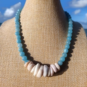 May include: A beaded necklace featuring light blue, round beads and a central section of irregular, light pink shell pieces. The necklace is displayed on a neutral-toned surface, with a blurred background of sky and water.
