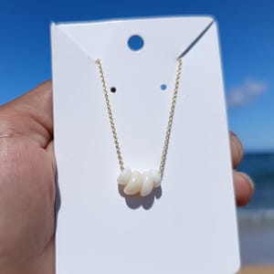 Peut inclure: Un collier délicat avec une chaîne dorée et cinq perles blanches de forme irrégulière. Le collier est présenté sur une carte blanche. Le fond montre un ciel bleu et la mer, créant une ambiance côtière.
