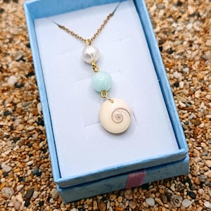 Pode incluir: Um colar dourado com uma pérola, uma conta azul clara e um pingente de concha creme com um design em espiral. O colar é exibido em uma caixa de joias azul clara, em um fundo de pequenas pedras.
