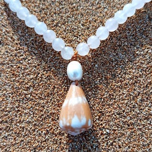 Puede incluir: Un collar con una cadena de cuentas blancas y redondas. Un colgante en forma de lágrima con patrones marrones y blancos cuelga debajo de una perla blanca. El collar se muestra sobre una superficie arenosa.