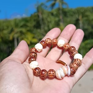 以下が含まれることがあります： 自然な要素を組み合わせた手作りのブレスレット。茶色とクリーム色のビーズ、貝殻が特徴です。ブレスレットは手で持たれており、背景にはヤシの木と青い空が広がる熱帯の風景が広がっています。