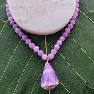 Puede incluir: Un collar de cuentas lavanda con un colgante en forma de lágrima. El collar está colocado sobre una piedra blanca y redonda. El colgante es de color púrpura claro con una base blanca y marrón. El fondo es una hoja verde.