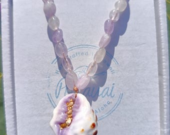 Puede incluir: Un collar hecho de cuentas de amatista lavanda con un colgante de concha. La concha es blanca con manchas marrones y tiene un interior morado. El collar se muestra en una caja blanca con el texto "Maui Sai Aloha" impreso en ella.