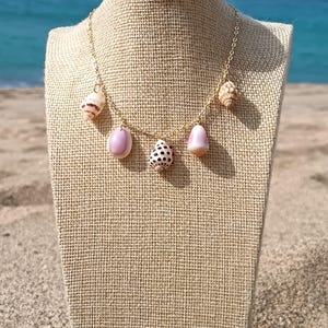 May include: A gold-colored chain necklace adorned with various seashells and pink pendants. The necklace is displayed on a burlap-covered bust, with a beach and ocean backdrop.