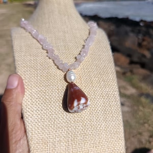 Puede incluir: Un collar con una cadena de cuentas de piedra rosa claro y un colgante. El colgante es una piedra en forma de lágrima con patrones marrones y blancos, coronada por una perla blanca. El collar se exhibe en un soporte beige.
