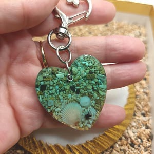 Könnte beinhalten: Herzförmiger Schlüsselanhänger mit silbernem Verschluss. Das Herz ist mit kleinen türkisfarbenen Steinen, Muscheln und klarem Harz gefüllt. Ein einzigartiges Accessoire mit Strand-Thema.