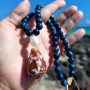 May include: A handcrafted necklace featuring a string of dark blue beads. The necklace has a large, pearly white pearl and a brown and white seashell pendant. The necklace is held up against a bright blue sky and ocean background.