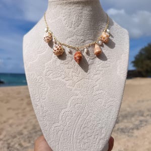 May include: A gold chain necklace featuring a selection of seashells and small white pearls. The seashells are in shades of tan, brown, and orange. The necklace is displayed on a cream-coloured jewellery bust against a beach backdrop.