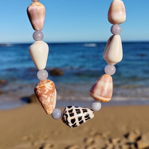 Puede incluir: Un collar con conchas marinas y cuentas de color púrpura claro. Las conchas son en tonos de blanco, beige y marrón, con una concha estampada en blanco y negro. El collar se muestra sobre un fondo de playa y océano.
