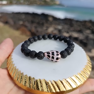 May include: A black lava bead bracelet with a seashell centerpiece. The bracelet is displayed on a white and gold decorative coaster, with a blurred ocean background.