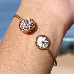 May include: Gold-toned cuff bracelet featuring two circular, shell-adorned accents. The shells display a brown and white striped pattern. The bracelet is a simple, open design, perfect for everyday wear. The bracelet is shown on a person's wrist.