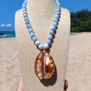 May include: A necklace featuring a single large, brown and white patterned cowrie shell pendant suspended from a strand of round, light blue beads. The beads are uniform in size and color, creating a smooth, continuous chain that leads to the shell.