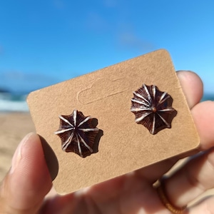 May include: A pair of brown and white stud earrings with a radial design, resembling a stylized flower or sunburst. The earrings are displayed on a small, rectangular, tan card. The background features a blue sky and ocean.