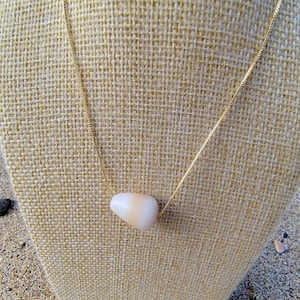 Op de afbeelding: Een delicate gouden ketting met een gladde, gebroken witte, druppelvormige hanger. De hanger hangt aan een fijne gouden ketting, wat een minimalistisch ontwerp creëert. De ketting wordt gepresenteerd op een neutraal gekleurde stof.