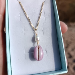 May include: A gold chain necklace featuring a white pearl and a pink seashell pendant. The necklace is displayed in a light blue gift box, indicating it's jewellery. The seashell has a natural, organic form.